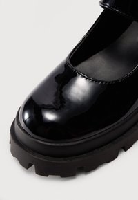 Svarte patent lær Mary Jane-sko med rund tå, tykk plattformsåle og justerbar stropp. Har teksturert grep på sålen.