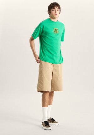 Grünes T-Shirt mit kurzen Ärmeln und Sonnenmotiv, kombiniert mit beigen Shorts. Weiße Socken und Leopardenmuster-Sneaker runden das Outfit ab.