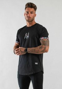 Svart t-shirt med blommönster på ärmarna och ett mönstrat "M"-logotyp på bröstet; har en rundad nederkant och ett etikett på nederdelen.