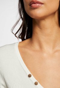 Haut en tricot côtelé blanc avec un col en V, présentant des détails de boutons dorés sur l'épaule et une teinte de peau lisse dans la zone visible.
