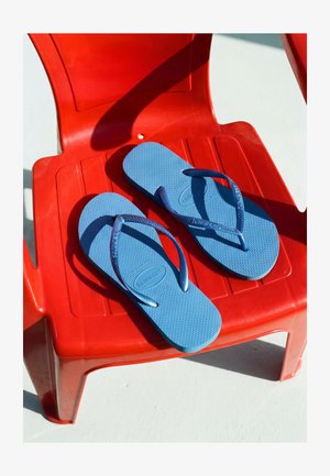 Paire de tongs bleues avec le nom de la marque sur les brides, placées sur une chaise en plastique rouge sous un fort soleil.