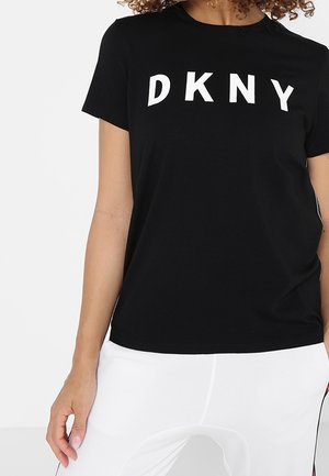 Kobieta w czarnej koszulce DKNY i białych spodniach, stojąca z lewym ramieniem swobodnie opuszczonym wzdłuż ciała na jednolitym tle.