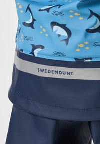 Gros plan sur un imperméable bleu avec des requins de dessin animé, de petits poissons jaunes et le texte "Swedemount" sur une bande réfléchissante grise.