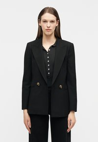 Γυναίκα με ίσια καστανά μαλλιά, φοράει μαύρο σακάκι με χρυσά κουμπιά πάνω από μαύρο πουκάμισο με κουμπιά, στέκεται μπροστά σε απλό λε υκό φόντο.