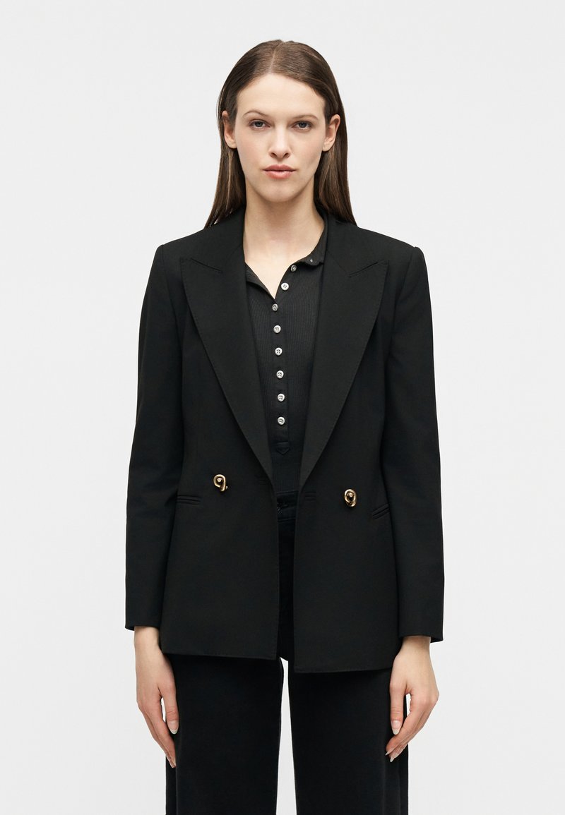 Γυναίκα με ίσια καστανά μαλλιά, φοράει μαύρο σακάκι με χρυσά κουμπιά πάνω από μαύρο πουκάμισο με κουμπιά, στέκεται μπροστά σε απλό λευκό φόντο.