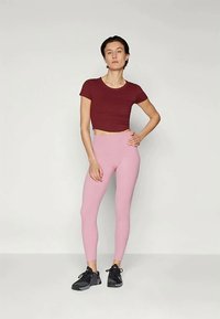 Burgundia lühike topp, millele on sobitatud heledat roosat retuusid, millel on vormiv disain ja sile tekstuur. Mudel kannab musti spordijalatsid.