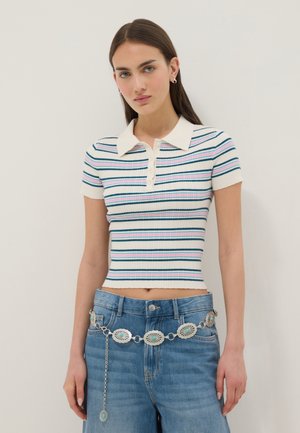 Giovane donna con capelli castani lisci che indossa una polo corta a righe e jeans blu con una cintura decorativa argentata con accenti turchesi.