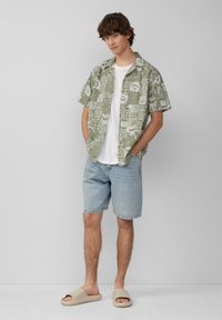 Chemise à manches courtes verte à motifs par-dessus un t-shirt blanc, associée à un short en denim bleu clair et des sandales à brides beiges.