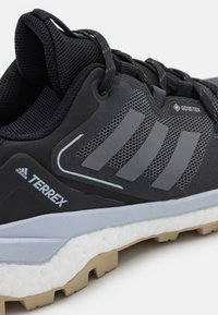 Černé trailové boty Adidas Terrex se šedými pruhy, štítkem Gore-Tex, bílou strukturovanou podrážkou a béžovým hrubým vzorkem podrážky.