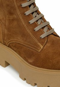 Brun ruskinds ankle boot med beige chunky sål. Har grå snørebånd, diskrete syninger og en tekstureret overflade.