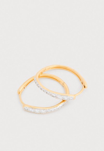 Złote kolczyki-koła z zakrzywionym wzorem i ukośną linią przejrzystych kamieni szlachetnych wzdłuż jednego brzegu. Gładkie metaliczne wykończenie.