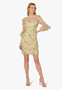 Vestito corto beige con silhouette attillata, caratterizzato da maniche trasparenti decorate con ricami floreali verdi e un sovrapposto semi-trasparente.