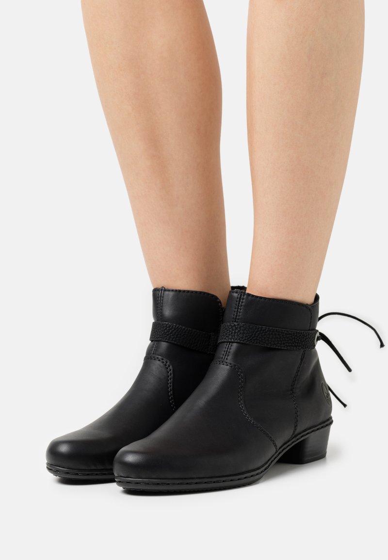 Bottines en cuir noir avec un petit talon, sangle texturée autour de la cheville et une finition lisse. Présente des détails de couture et un accent en lacet.