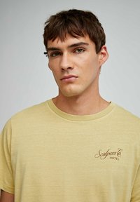 Lys gul bomulds T-shirt med rund hals, med et brunt "Scalpers Co. HOTEL" logo på venstre bryst. Blød tekstur, afslappet pasform.