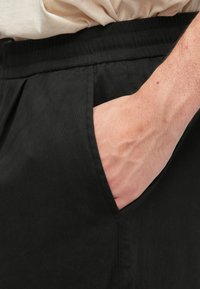 Pantalon noir à texture lisse, avec poches latérales et ceinture élastique. L'image montre une main reposant dans la poche.