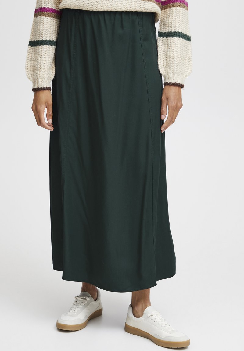 Jupe de couleur vert foncé, arrivant aux genoux, en tissu lisse, avec une taille élastique et des détails de couture sur les côtés, assortie à des baskets blanches.