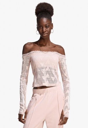 Giovane donna nera con acconciatura afro che indossa un top in pizzo rosa pallido con maniche lunghe e scollatura a spalle scoperte e pantaloni rosa chiaro abbinati, in posa.