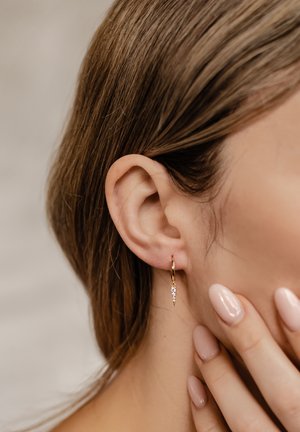 Gold hoop earring features a dangling, pointed pendant adorned with small clear stones. Textured smooth surface, subtle shine, minimal design.