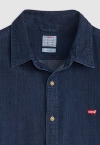 Denimska košulja tamnoplave boje s oštrim ovratnikom, zlatnim gumbima i crvenom Levi's etiketom na lijevoj strani prsiju. Ima uski kroj.