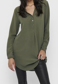 Chemise à manches longues de couleur vert olive en tissu léger. Elle présente une patte de boutonnage, un ourlet arrondi et des manches à revers, portée sur un legging noir.