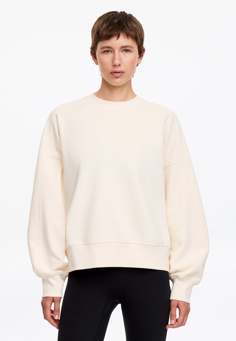 Jeune personne aux cheveux bruns courts portant un sweat-shirt crème ample et un pantalon noir, debout devant un fond blanc uni.