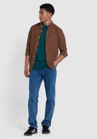 Bruine knopshirt over een groene T-shirt, gecombineerd met blauwe spijkerbroeken en zwarte sneakers met witte accenten. Model staat tegen een neutrale achtergrond.