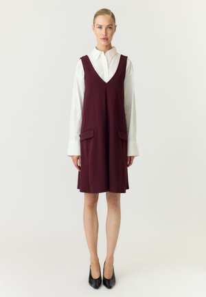 Femme portant une chemise blanche à manches longues sous une robe bordeaux sans manches longueur genou avec poches à rabat, associée à des talons noirs.