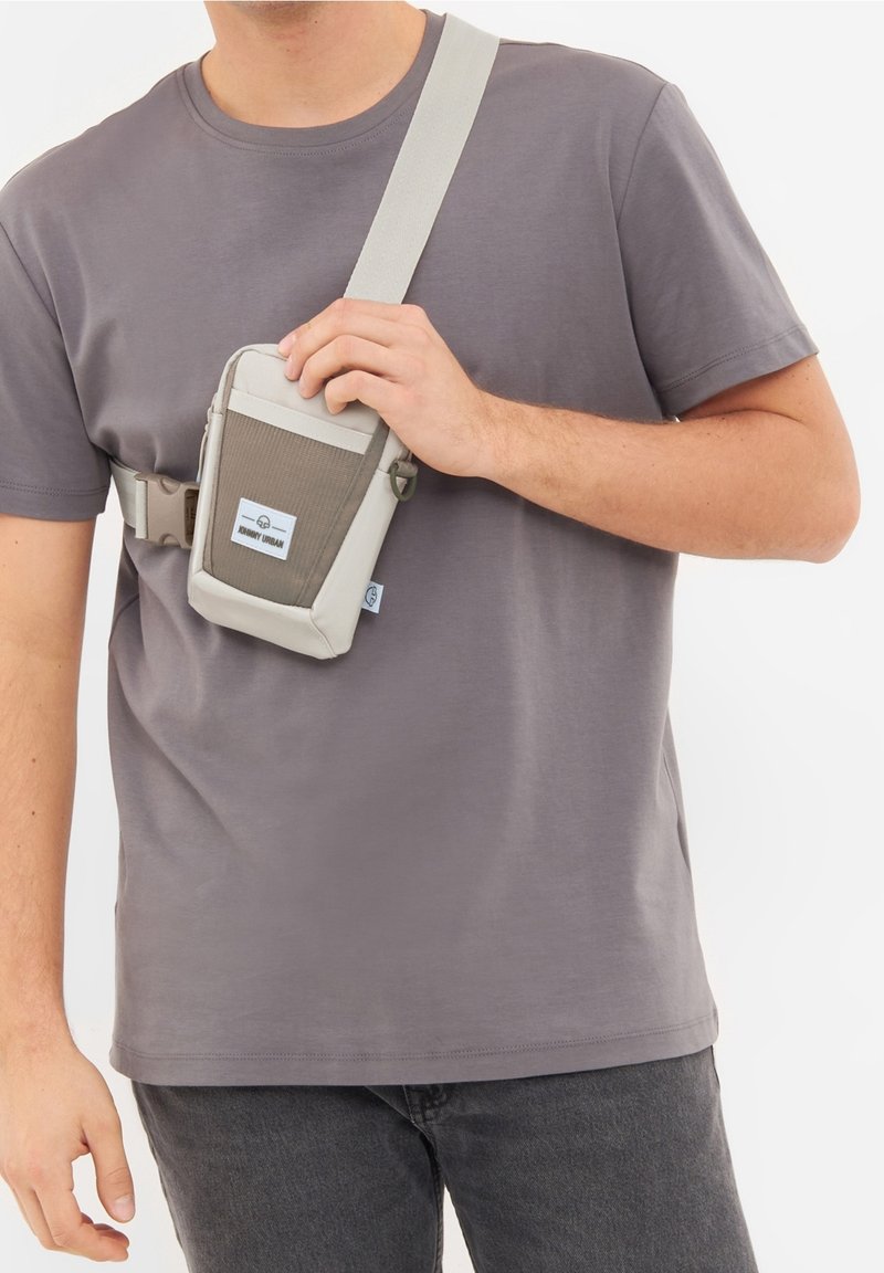 Šedá kabelka přes rameno s texturovaným šedým předním panelem, béžovými akcenty, nastavitelným popruhem a nášivkou s logem. Kompaktní design vhodný pro nezbytnosti.