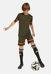 Dunkelgrünes Fußballtrikot und Shorts mit orangefarbenen Akzenten, kombiniert mit schwarzen Socken mit weißen Streifen und einem Fußball unter dem Fuß.