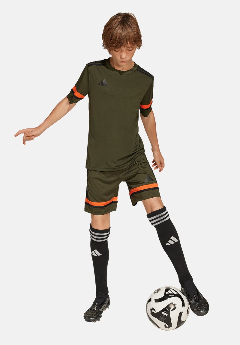 Dunkelgrünes Fußballtrikot und Shorts mit orangefarbenen Akzenten, kombiniert mit schwarzen Socken mit weißen Streifen und einem Fußball unter dem Fuß.
