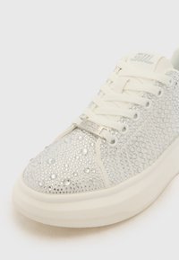 Baskets blanches avec surface texturée, ornées de strass en argent. Présentent des lacets plats et une semelle épaisse et texturée. Détail de marque en argent.