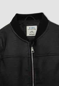 Czarna bomberka z syntetycznej skóry z prążkowanym kołnierzem i zapięciem na zamek. Metka wskazuje na "KOLEKCJĘ DZIECIĘCĄ" w rozmiarze 7-8 lat, marka DeFacto.