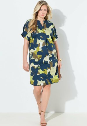 Donna che cammina con un vestito corto al ginocchio a maniche corte con motivo floreale astratto verde, blu e marrone, indossa sandali e tiene una piccola borsa.