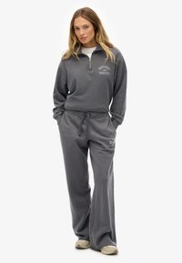Frau steht in einem dunkelgrauen Superdry Athletics Zip-Up-Sweatshirt und passenden weit geschnittenen Sweatpants mit beigen Sneakers vor einem weißen Hintergrund.