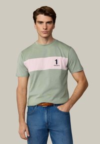 Ung mand iført salviegrøn T-shirt med bred, lys pink vandret stribe og nummer 1 Hackett-logo, blå jeans og brunt bælte.