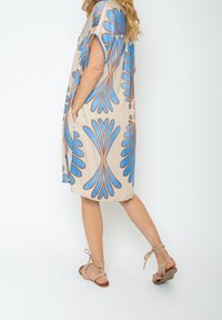 Vestido beige con grandes patrones florales azules y acentos marrones. Corte holgado con mangas cortas y bolsillos laterales. Llevado con sandalias beige de cordones.