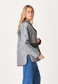 Grå quiltet jakke med bælte i taljen, høj krave, lange ærmer og et struktureret mønster. Accentueret med sort kant.
