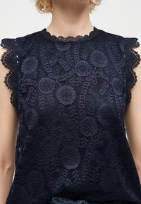 Κοντινό πλάνο πάνω από μπλε σκούρο δαντελένιο τοπ με λουλουδάτα σχέδια, κυματιστές άκρες στα κοντά μανίκια και στρογγυλή λαιμόκοψη σε άτομο με σγουρά μαλλιά.