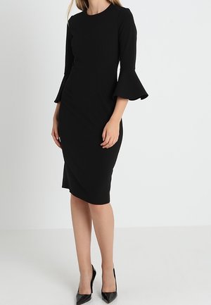 Robe noire ajustée avec un col rond, des manches longues et des poignets évasés. Fabriquée en tissu lisse. Portée avec des talons hauts noirs.