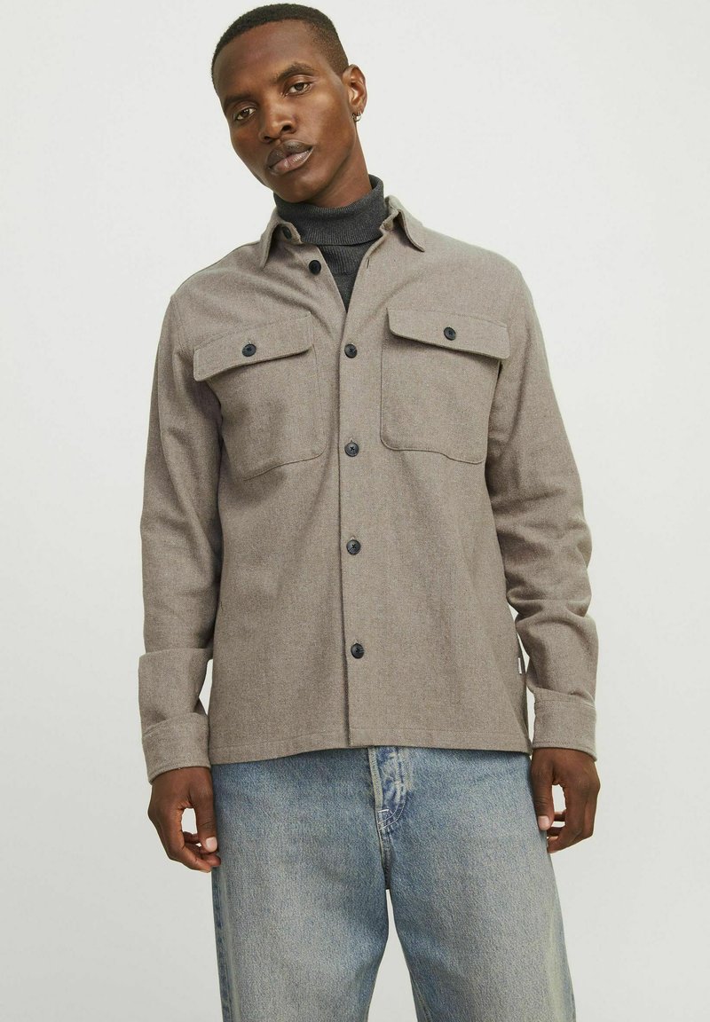 Camicia grigia con bottoni, due tasche sul petto, realizzata in tessuto morbido. Presenta bottoni neri e un colletto. Abbinata a jeans chiari.
