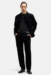 Czarna kurtka z sztruksu noszona na czarnej koszuli, zestawiona z czarnymi sztruksowymi spodniami i błyszczącymi czarnymi butami. Minimalistyczny design i jednolite kolory.