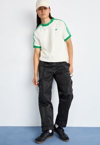 T-shirt branca com riscas verdes e logótipo, calças cargo pretas com bolsos laterais e ténis pretos. Roupa casual com acentuados toques desportivos.