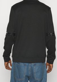 Felpa nera con materiale testurizzato, caratterizzata da strisce nere lucide sulle maniche e un collo e orlo a coste. Indossata con jeans blu.