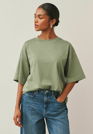 Frau mit Haaren im Dutt trägt ein grünes übergroßes T-Shirt und blaue Jeans, steht mit einer Hand in der Tasche vor einem einfarbigen Hintergrund.