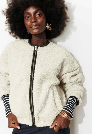 Femme avec coiffure afro portant une veste zippée blanche texturée sur un t-shirt à manches longues rayé noir et blanc, boucles d'oreilles en or et bracelets.