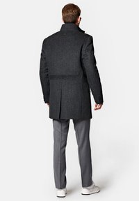 Czarny wełniany płaszcz z wysokim kołnierzem, pojedynczym rozcięciem i zapinanymi mankietami, w zestawie z jasnoszarymi spodniami i białymi trampkami.