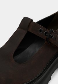 Chaussure en cuir marron avec un design à barrette T, sangle avec boucle et surface texturée. Bout rond avec une semelle en caoutchouc noir.