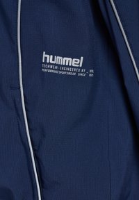 Marineblauwe sportkledingstof met wit "hummel"-logo en tekst "Techwear engineered by HML Performance Sportswear since 1923" met witte biezen.