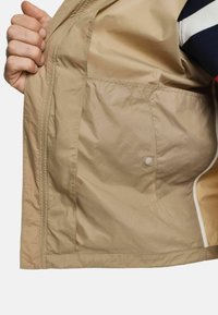 Giacca leggera beige con interni che mostrano una tasca laterale con bottone a scatto, texture liscia e bordatura a contrasto crema lungo il bordo.