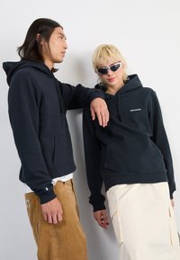 Tume sinine kapuutsiga särk, millel on esikotid ja nöörid, koos beeži sametist pükste ja heledat värvi seelikuga. Mudel kannab prille.